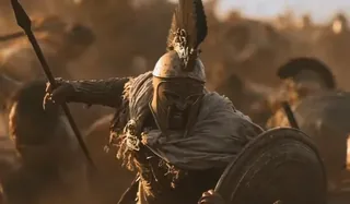 A Spartan warrior in bronze armor charges forward with a spear and shield on a dusty battlefield during a golden sunset.