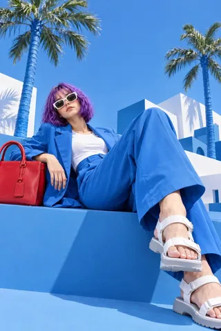 Full-body low-angle fashion photograph of a character in a cobalt-blue suit lounging on a blue architectural ledge under bright summer sunlight.