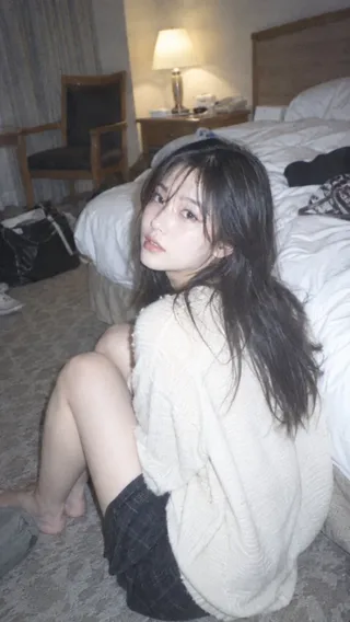 A young Japanese woman with long black hair, wearing an ivory knit top, sits casually on a carpet, looking back at the camera with a relaxed expression, evoking a vintage 2000s digital snapshot aesthetic.