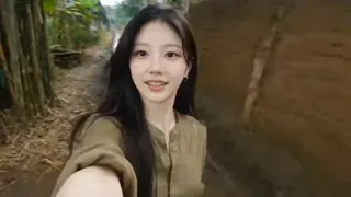 Authentic Southeast Asian village scene with a woman in a damp, oversized shirt and burgundy trousers, filmed with a shaky, lo-fi flip cam aesthetic, showing rural life.