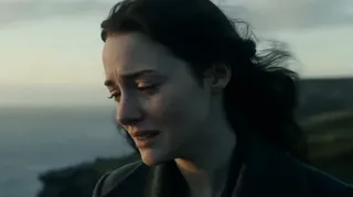 A woman stands on a jagged cliff, her coat being torn by the wind, overlooking a powerful ocean with waves crashing against the rocks below. The camera perspective shifts to an aerial view, showing her as a lone figure against the vast sea.