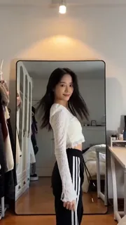 A young East Asian woman with long black hair, wearing a white sweater and athletic pants, dancing in front of a bedroom mirror.