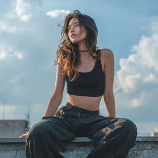 A full-body editorial portrait of a young East Asian woman in a black crop top and cargo pants sitting on a rooftop ledge during golden hour.