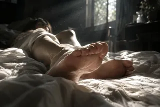 Close-up of feet on a white sheet, illuminated by dappled sunlight filtering through leaves, with a strong contrast of light and shadow and a grainy film texture.