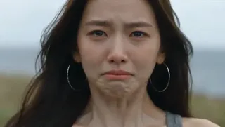 Young East Asian woman in a grey tank top, with large silver hoop earrings, suppresses tears, then pulls her shirt up to cover her mouth and nose, before letting it fall, revealing a weary face.