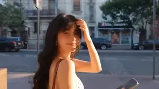 A woman smiles at the camera while walking on a beach during golden hour, with a handheld vlog-style camera following her.
