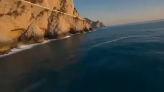 Continuous aerial drone footage of a dramatic coastline with cliffs, a winding road, a village, and sailboats, bathed in golden hour light.