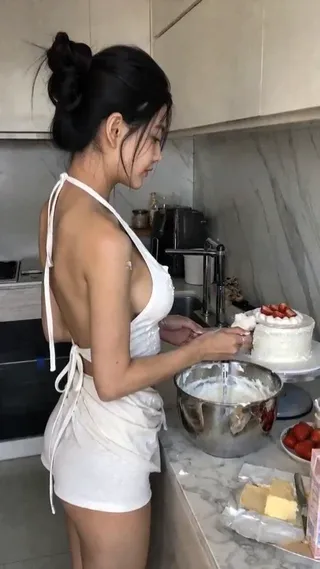 A young Asian woman with a black bun, wearing a white halter top and light pants, playfully eats a strawberry cake in front of a marble counter, looking at the camera with a smile and blinking.