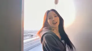 Korean female idol Hana, with long dark hair, smiles at a consumer camcorder on a hidden rooftop, overlooking a city skyline at sunset.