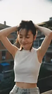 A young Chinese woman with a casual outfit and natural makeup sits on a low wall outside a home, adjusting her hair in a narrow alleyway with red brick buildings and trees.