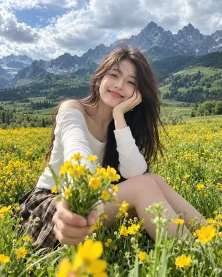 A smiling young woman in a white top and plaid skirt sitting in a vibrant yellow wildflower field with dramatic green mountains in the background.
