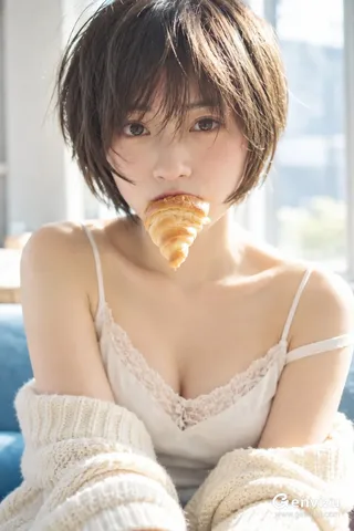 A vertical portrait of an East Asian woman with a short dark bob, holding a small croissant by her lips while sitting by a bright window in an ivory camisole.
