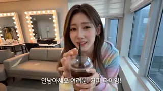 A smiling Korean female idol in a pastel outfit sits in a cozy dressing room, holding an iced coffee and her phone, decorating her cup with a sticker.