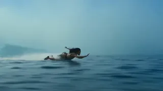 A young woman with wind-blown hair, a determined expression, rides a board inches above a misty ocean with large waves crashing behind her.