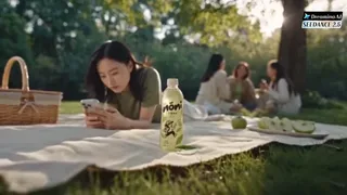 A young Chinese woman at a park picnic, initially engrossed in her phone, then picks up a Nōni green tea bottle. A small animated character detaches from the bottle to playfully guide her attention back to the present moment and her friends.