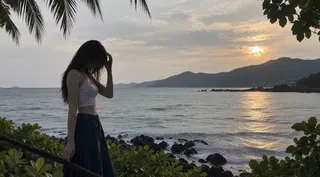 A solitary young woman with long dark hair standing by a rope railing on a rocky tropical shoreline during a warm golden hour sunset.