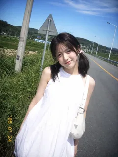 A young East Asian woman with dark hair in low pigtails, wearing a white dress, stands on a country road with a slight smile, looking at the camera, in a vintage CCD photo style.