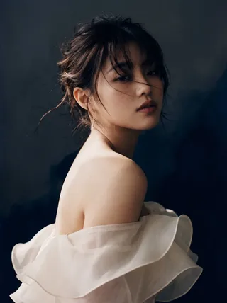 High fashion editorial portrait of a young woman with dark hair, wearing an ivory silk organza ruffled gown against a grey and blue ink-splatter studio wall.