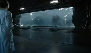 A lone female researcher in a white lab coat stands before a massive reinforced glass wall in a futuristic laboratory, facing three colossal, terrifying dragons emerging from a haz