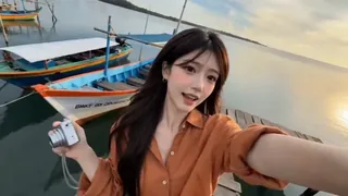 Authentic Indonesian woman in a terracotta shirt and burgundy pants, recording a selfie vlog in a coastal village, with fishing boats and a stray dog visible, showcasing late-2000s