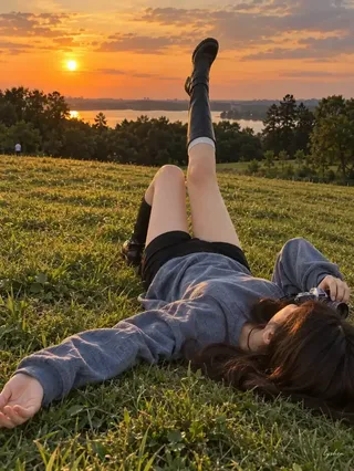 An East Asian woman lying in a grassy field during a warm golden sunset, holding a camera, with a distant treeline and sky.