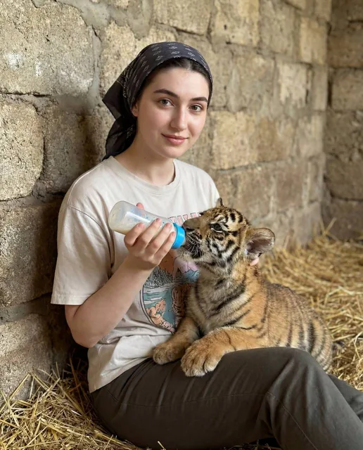 A young woman with brown hair in a bun and a patterned headscarf, wearing a graphic t-shirt, smiles gently as she holds a baby bottle to feed a tiger cub lying on the floor in a ru