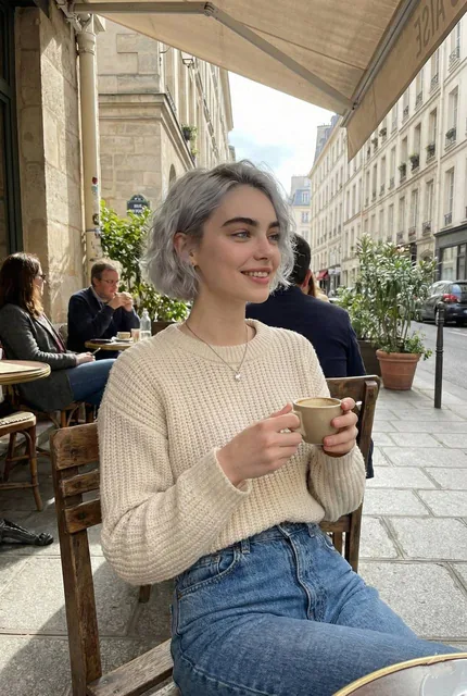 A candid lifestyle portrait of a young woman sitting at an outdoor Parisian café, holding a ceramic coffee cup with both