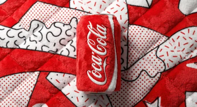 A beverage can branded [BRAND NAME], entirely made from vibrant, fluffy plush fabric, positioned centrally against a sof