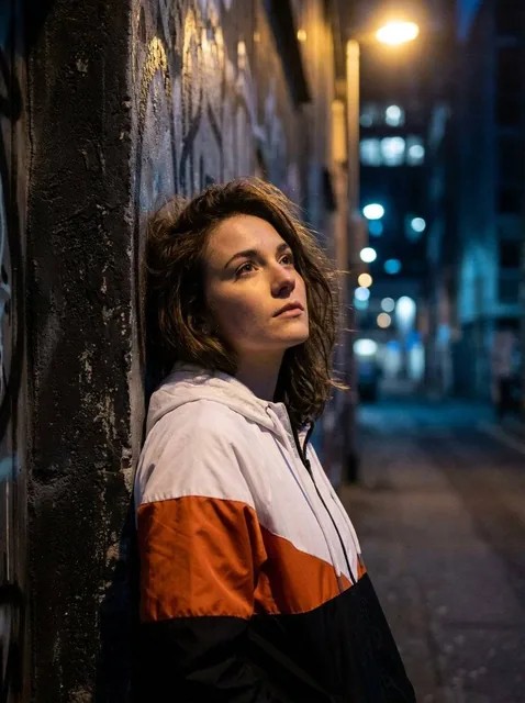 A young women with tousled brown hair, wearing a white, orange, and black color-block jacket, leans against a dark pilla