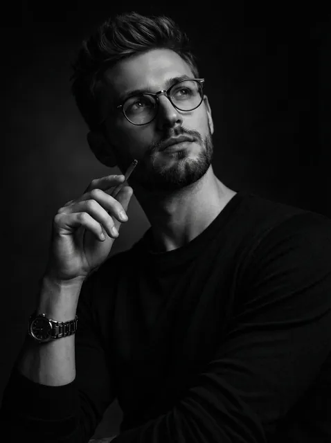A dramatic black and white studio portrait of a thoughtful man with short styled hair, light stubble beard, and round ey