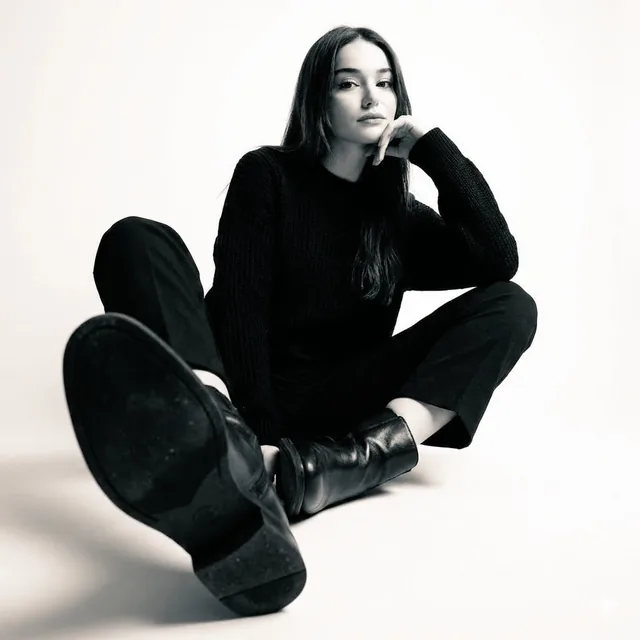 High-contrast black-and-white studio photo of a woman seated on the floor wearing a black sweater, black trousers, and b