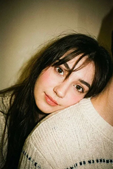 Close-up, high-angle selfie of a young woman with long black hair, wearing a cream knit sweater, leaning on her partner's shoulder in a warm home interior, captured with a disposab