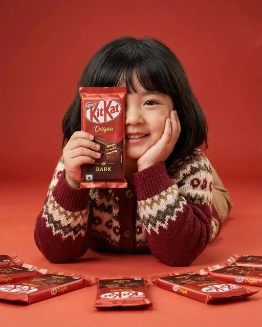 A cute young girl with short black hair and bangs smiling while holding a red KitKat Dark chocolate bar covering one eye