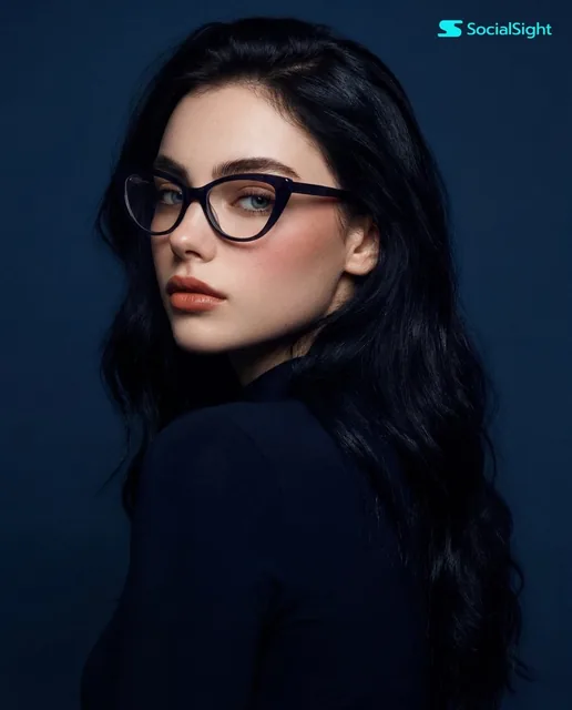 A young woman with long, dark hair and striking cat-eye glasses is captured in a studio portrait against a solid blue ba