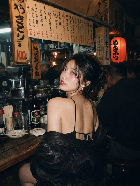 A beautiful young East Asian woman with long dark wavy hair and striking makeup, sitting at a wooden counter in a cozy,