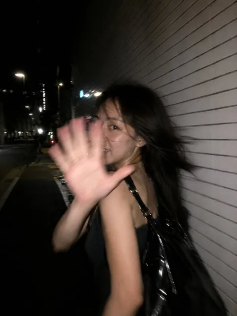 A young East Asian woman in a black dress is caught off guard by a camera at night, raising her hand to block the shot as she glances back with a playful smile, captured with stron
