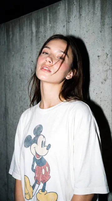 A high-resolution, close-up editorial-style portrait of a young woman standing against a textured gray concrete wall, ca