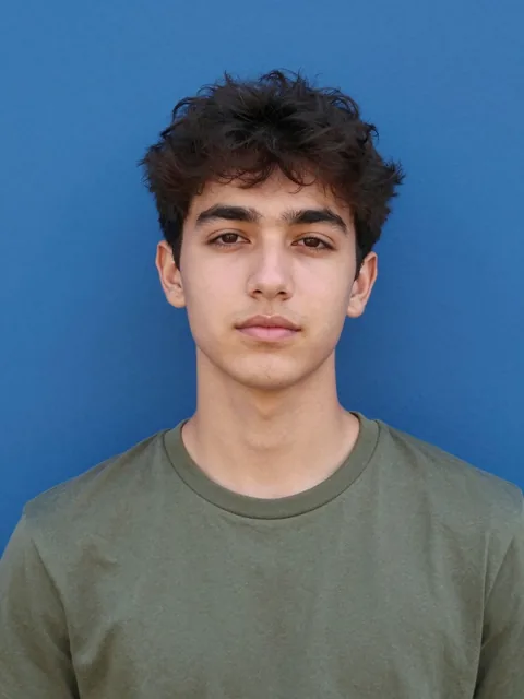A young man with dark, textured hair poses for a centered portrait against a flat, vibrant ultramarine blue wall. The li