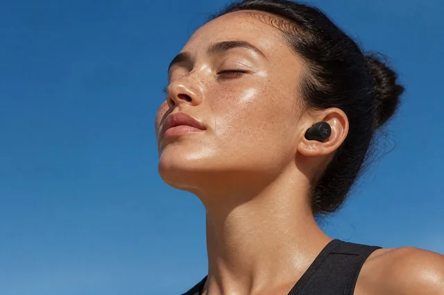 A serene close-up of a freckled woman with wireless earbuds, eyes closed, against a clear blue sky. Perfect for tech or