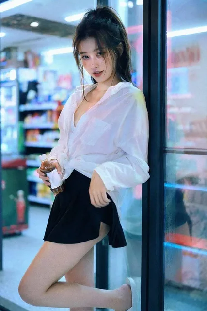 A young Chinese woman with dark hair in a messy ponytail, wearing an oversized white shirt and mini skirt, leans seductively against a convenience store glass door at night, illumi