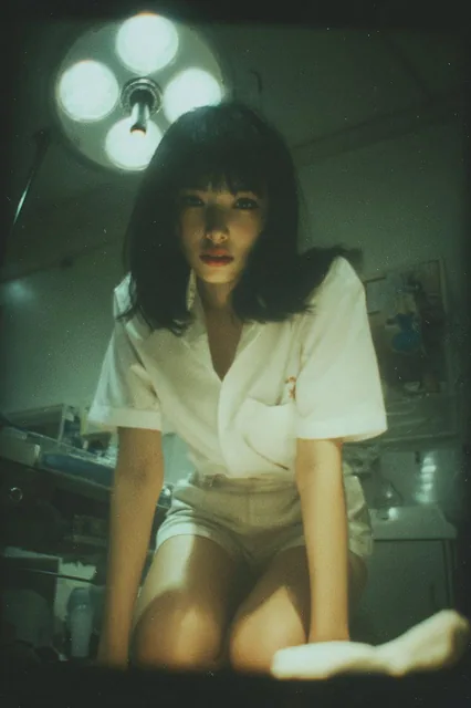 A beautiful Japanese woman with black hair and bangs, wearing a white shirt and shorts, kneels on an operating room bed while cooking rice, with a relaxed expression under soft overhead lighting in a retro Polaroid photo with grainy texture and warm tones.