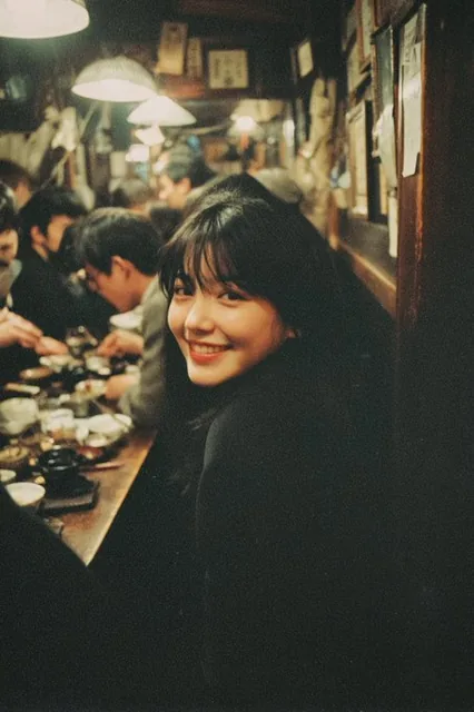 A candid photo of an Asian woman with black hair smiling while sitting at a dinner table in Tokyo's dimly lit bar district during a New Year's Eve party, captured with a Polaroid camera featuring a grainy texture and in-focus style.