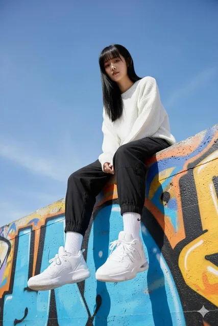 Low-angle view of a stylish young woman sitting on a graffiti-covered wall, wearing an oversized white sweater and black joggers, with a focus on her detailed Jordan sneakers.
