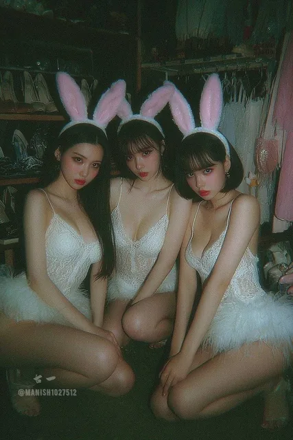 Three young Asian women in pink bunny ear headbands and white lace bodysuits posing seriously in a dim cluttered bedroom closet with warm lighting, grainy texture, and a 1990s film aesthetic.