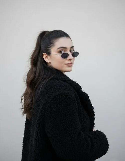 A woman with long dark wavy hair in a high ponytail, wearing small oval sunglasses and a black fluffy jacket, looking over her shoulder with a slight smirk against a plain light-colored wall.