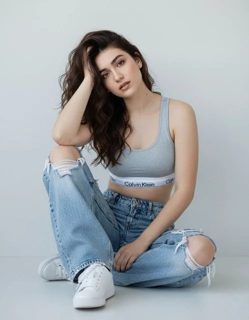 A woman in a light-gray athletic crop top with thick straps and Calvin Klein band, wearing extremely baggy distressed light-wash jeans with heavy rips at knees and thighs, seated on the floor against a bright plain background with legs bent and crossed, right arm up with hand on head, direct intense