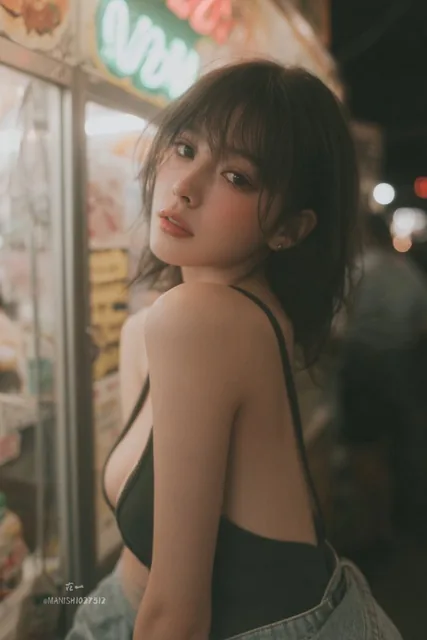 A stealthy candid photo of an Asian woman with straight hair and bangs, rosy cheeks, wearing a deep-neck tank top and jeans, browsing stalls with a curious gaze in low-light conditions with blurred neon signs and grainy texture.