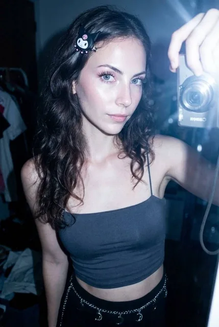 A teenage girl with a dark brown wavy hair and a Sanrio Kuromi hair clip, wearing a dark grey spaghetti strap crop top and a silver chain belt with moon charms. Her face has bright eyes with blue-grey contact lenses, fair skin with a rosy glow, and light pink makeup. The image is taken with a Canon