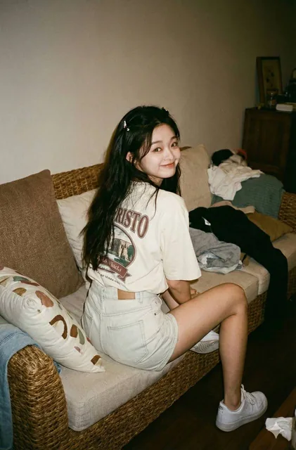 A hyperrealistic vintage lifestyle photo of a woman sitting on a woven sofa with her back turned, looking over her shoulder with a playful expression, in a cozy messy room with warm dim lighting and soft film grain.
