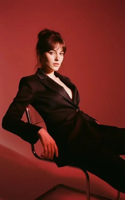 A high-contrast black and white editorial portrait featuring a woman in a ribbed black turtleneck top, with messy wind-blown updo, wispy bangs, and stray hair framing her face. She has high cheekbones, subtle smoky eye makeup, and natural lip color, rendered in monochrome with sharp focus on the eye
